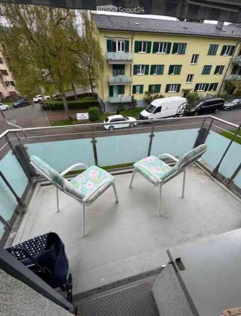 Balcony with two lounge chairs and a small table, overlooking a street with parked cars and neighboring buildings.