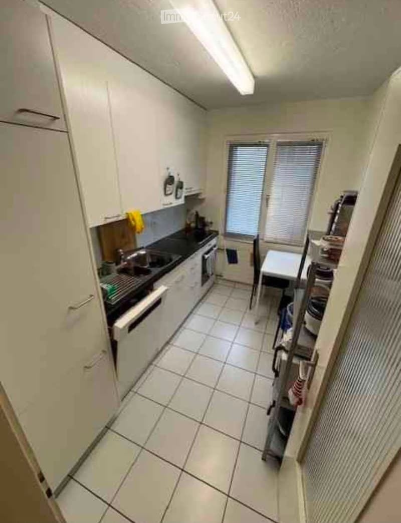 Modern kitchen with white cabinets, tiled floor, sink, oven, dishwasher, and a small table with a chair near the window.