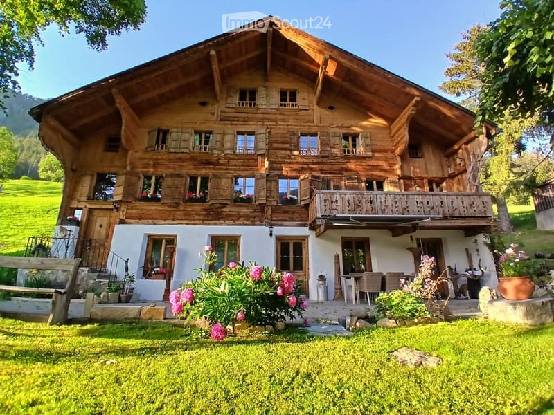 A two-story wooden house with a balcony, surrounded by a well-maintained garden with flowers and a bench.