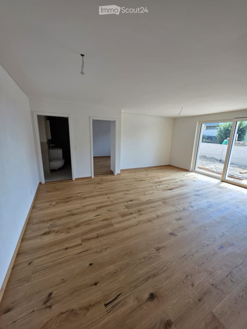 Empty room with light wooden flooring, white walls, and large sliding glass door leading to an outdoor area. Two doorways lead to adjacent rooms, one of which contains a toilet.