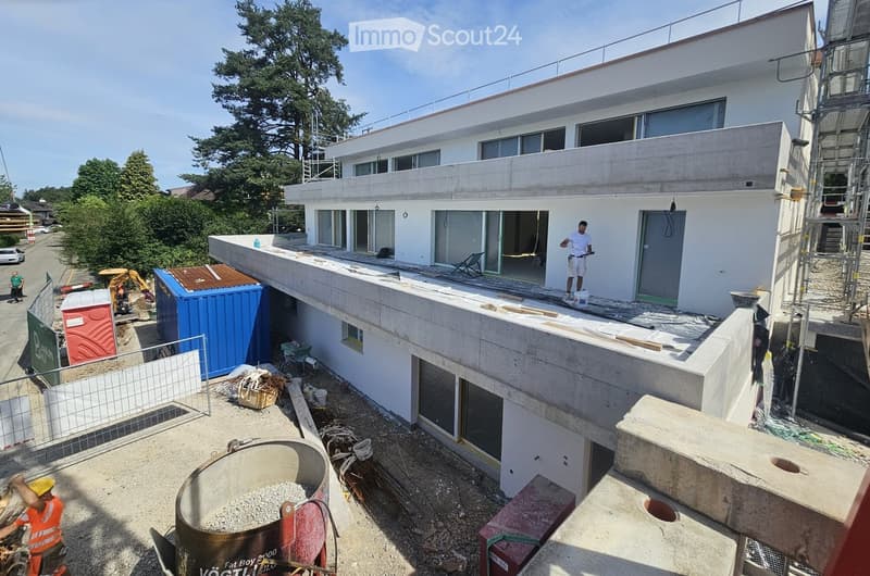 Modern two-story building under construction with large windows, a flat roof, and a balcony area. Workers and construction materials are visible around the site.