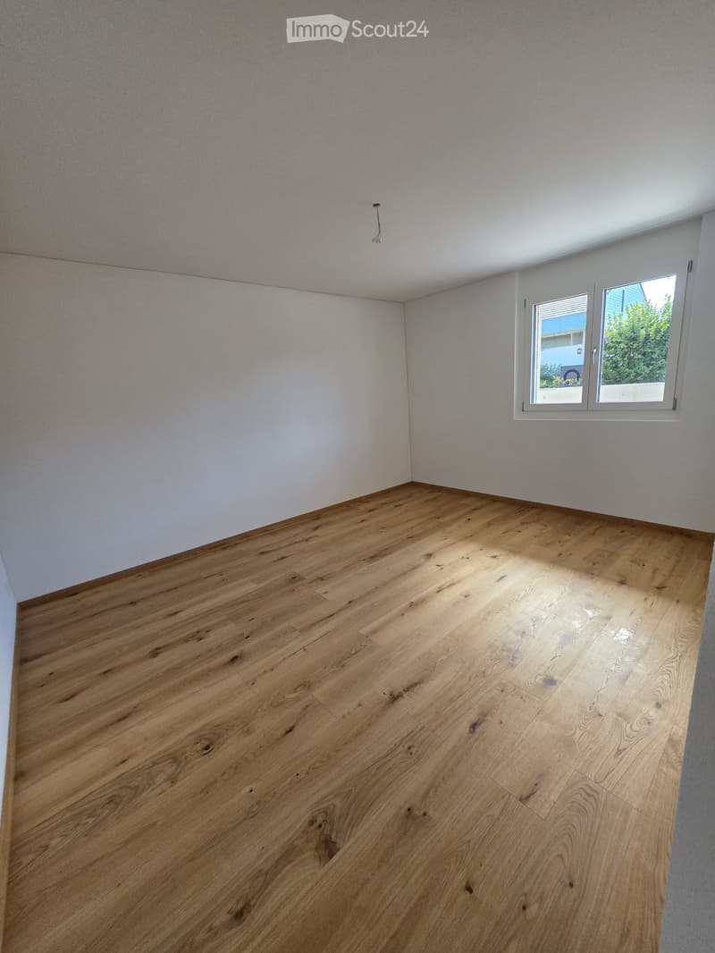 Empty room with wooden flooring and white walls, featuring a window with a view of greenery outside.
