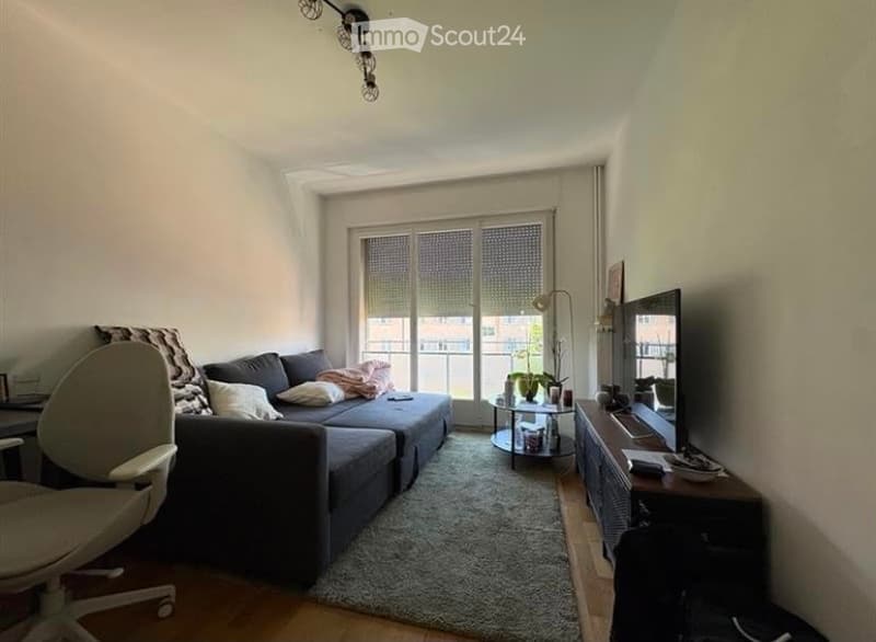 Modern living room with a grey sectional sofa, coffee table, TV stand, floor lamp, and a small round table near the balcony. The room features large windows with blinds and a cozy rug on the floor.