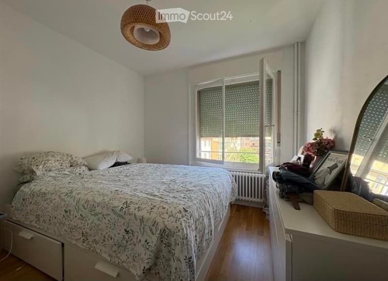 Cozy bedroom with a large bed, nightstand, mirror, wicker basket, and a window with blinds. The room features wooden flooring and white walls.