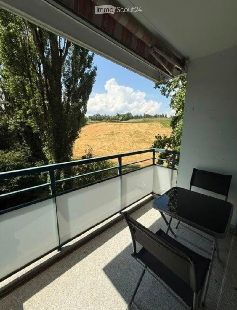 Balcony with a table and chair, overlooking a field with trees and a clear sky.