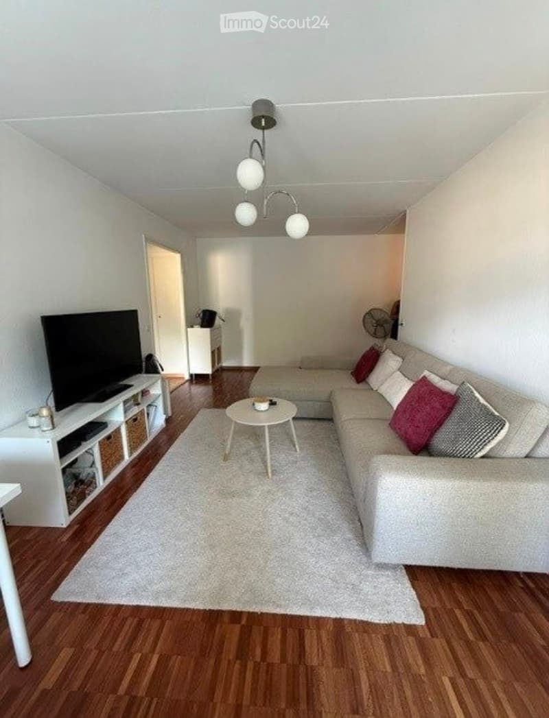 Modern living room with light-colored sofa, coffee table, TV, side table, and fan. Wooden flooring and white walls.