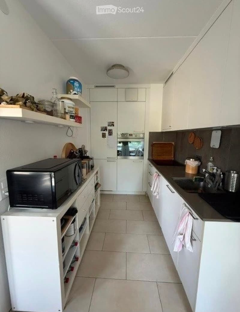 Modern kitchen with white cabinets, tiled floor, microwave, oven, sink, and shelves with various items.