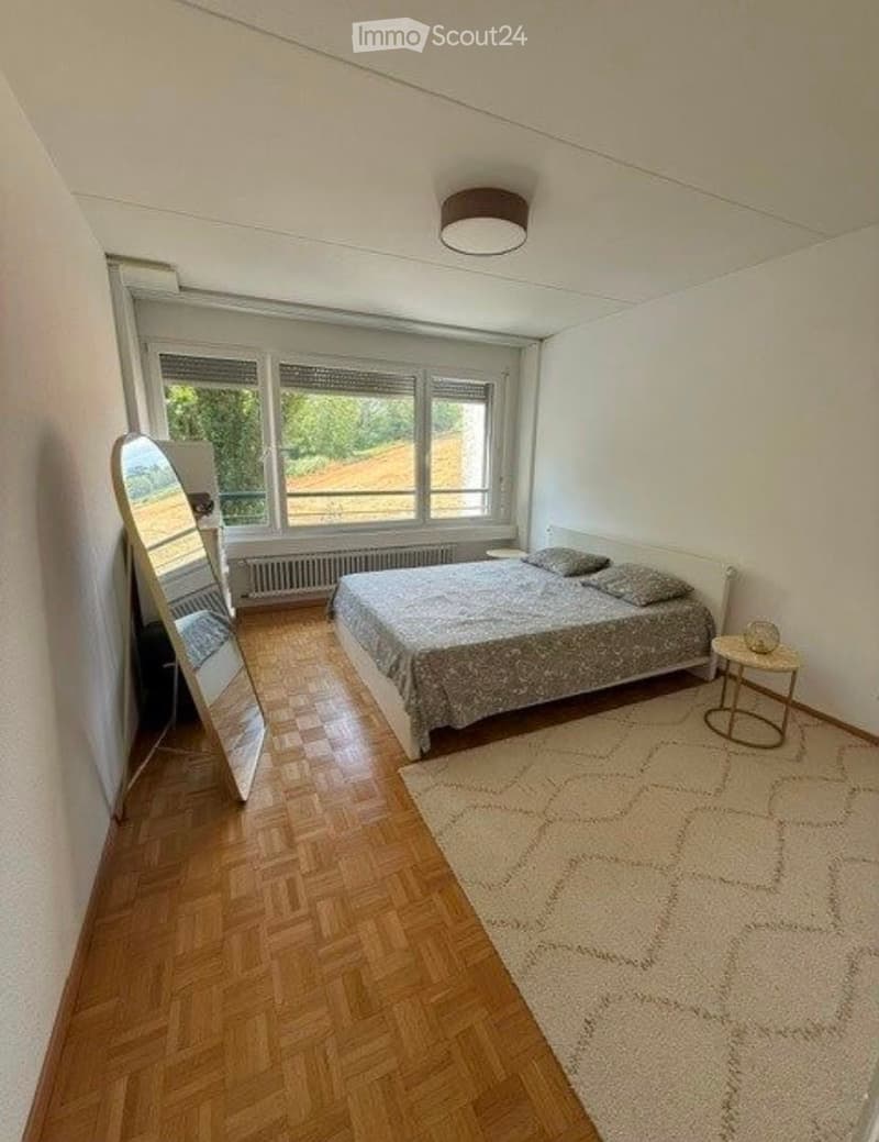 Bright bedroom with wooden flooring, large windows, a bed with patterned bedding, a mirror, a nightstand, and a small table with a decorative item.