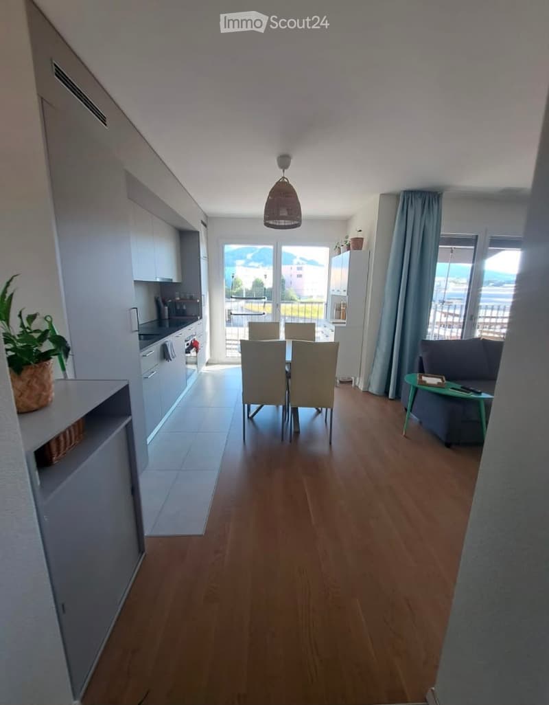 Open-plan living and dining area with wooden flooring, a grey sofa, a small green coffee table, a dining table with four chairs, and a modern kitchen with white cabinets and a refrigerator. Large windows offer a scenic mountain view.