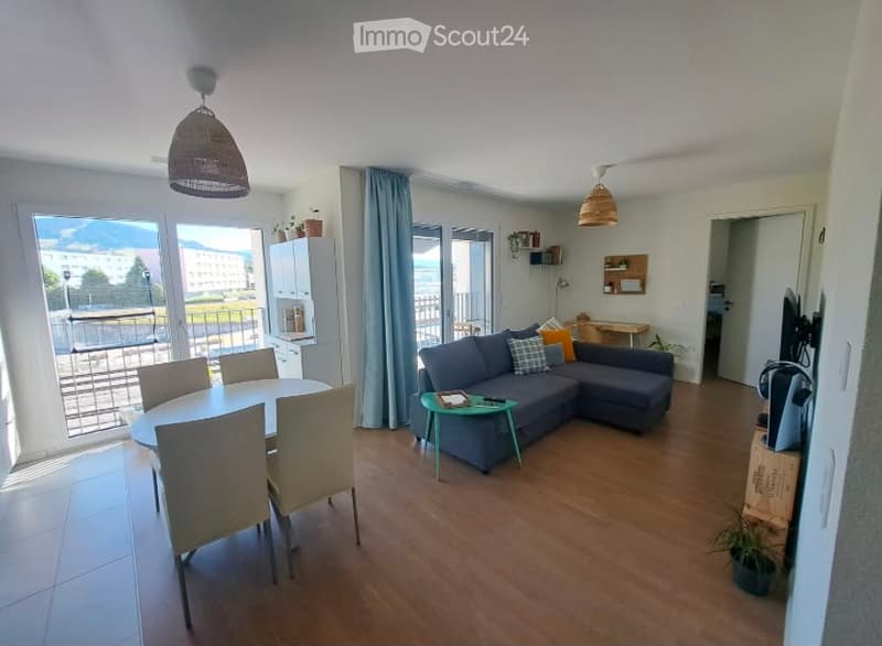 Open-plan living and dining area with wooden flooring, large windows, and a balcony. Features a grey sectional sofa, coffee table, dining table with chairs, and pendant lights.