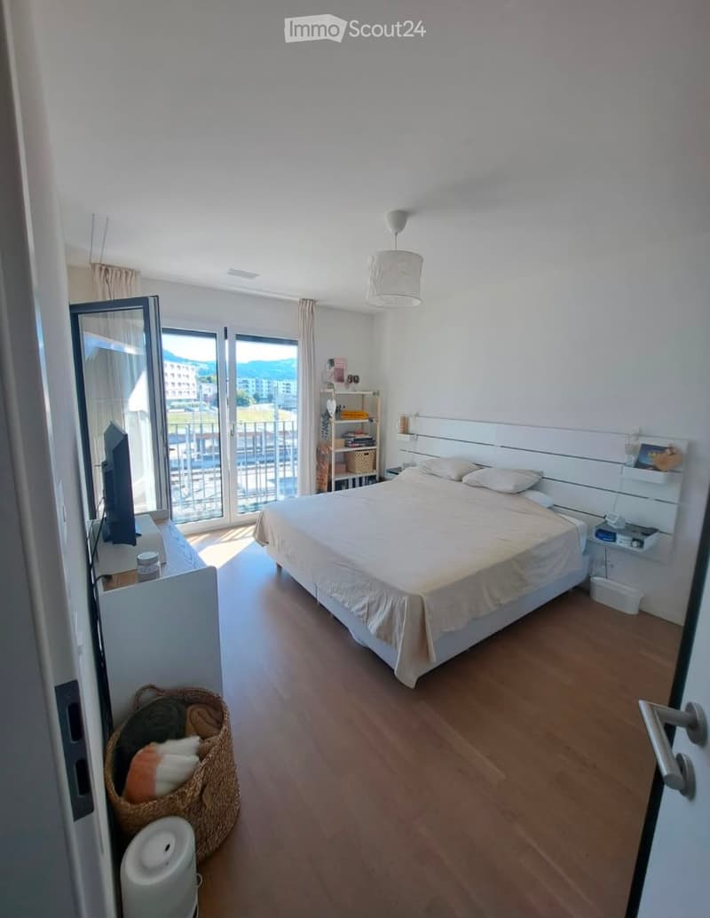 Modern bedroom with wooden flooring, large bed, nightstand, mirror, wicker basket, and a view of the city from the balcony.