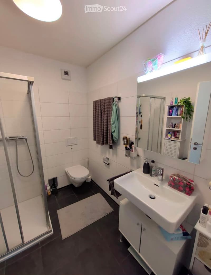 Modern bathroom with white tiles, a toilet, a sink with a mirror, a glass-enclosed shower, and a towel rack with towels. The room features a sleek design with a black floor and a small cabinet under the sink.