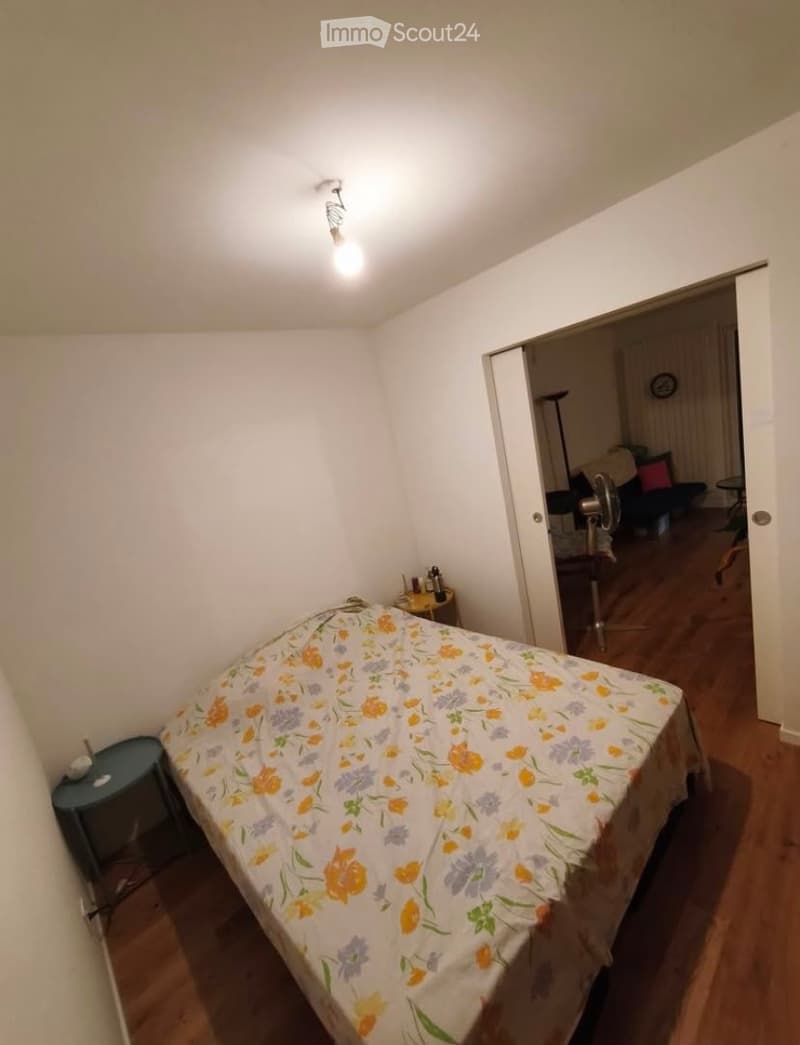 Bedroom with a single bed covered in a floral-patterned bedspread, a small nightstand, and a fan visible in the adjacent living area.