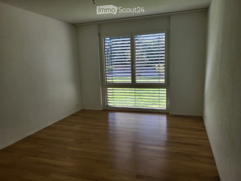 Empty room with wooden flooring and large window with blinds, showing a view of greenery outside.
