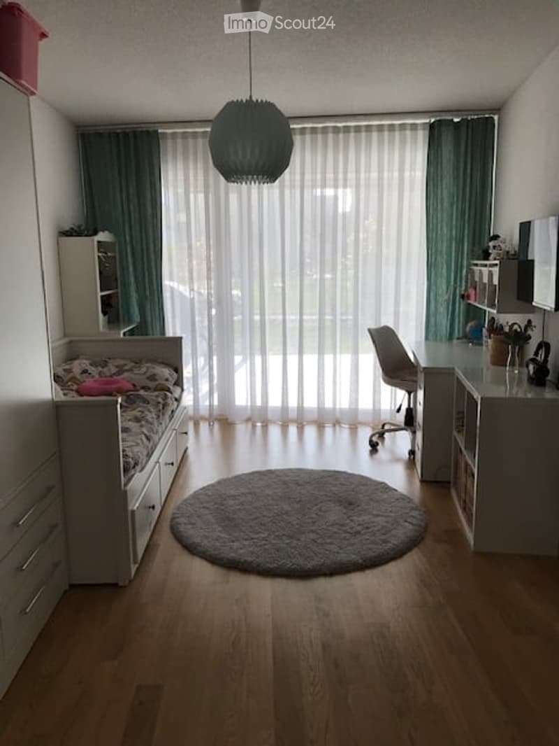 A modern, well-lit bedroom with a bed, desk, chair, wardrobe, and large window with curtains. The room features a round rug and a stylish hanging lamp.