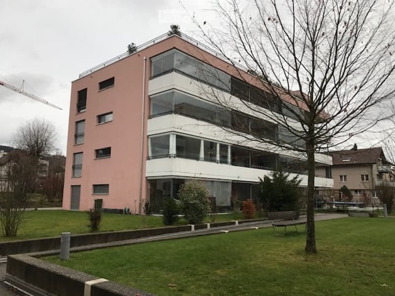 Modern multi-story building with pink facade and large glass windows, surrounded by a grassy area with trees and benches.