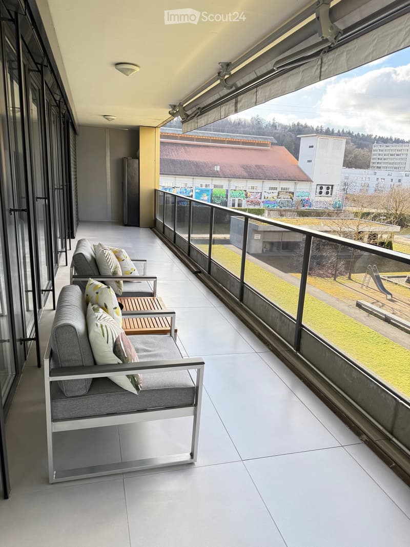 A balcony with a metal railing and large sliding glass doors, modern furniture with decorative pillows, tile flooring, and a scenic city view.