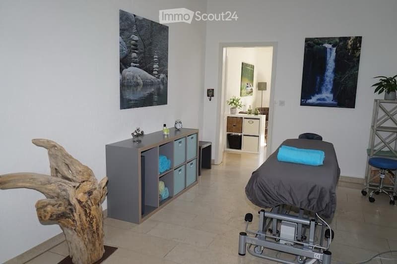 A well-lit, modern room with a massage table, storage cabinet, decorative wooden sculpture, picture frames, and a small plant. The space is clean and organized, suitable for relaxation or therapy.
