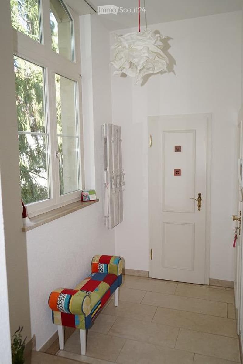 A brightly colored bench sits against a white wall in a hallway with large windows and a white door. A mirror and a decorative chandelier are also visible.