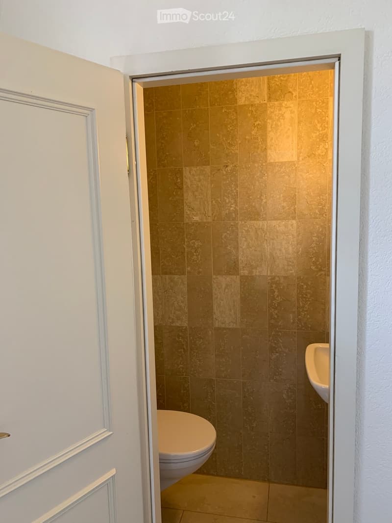 A small bathroom with beige tiled walls and floor, featuring a toilet and a sink visible through an open door.