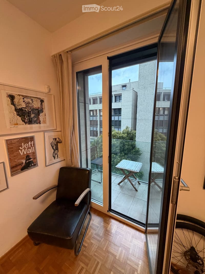 Modern room with wooden flooring, large sliding glass doors leading to a balcony, a black leather chair, and framed artwork on the wall.