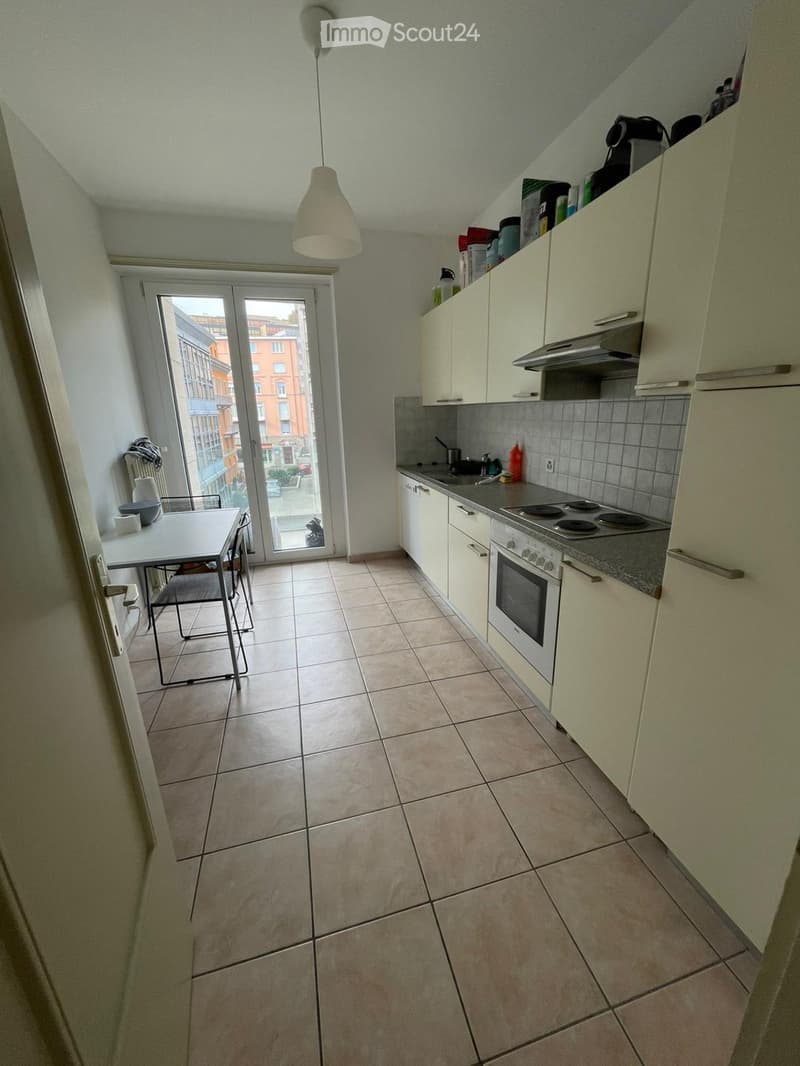 Modern kitchen with white cabinets, a four-burner stove, an oven, and a dining area with a table and chairs. The space features tiled flooring and a large window offering a view of the city.