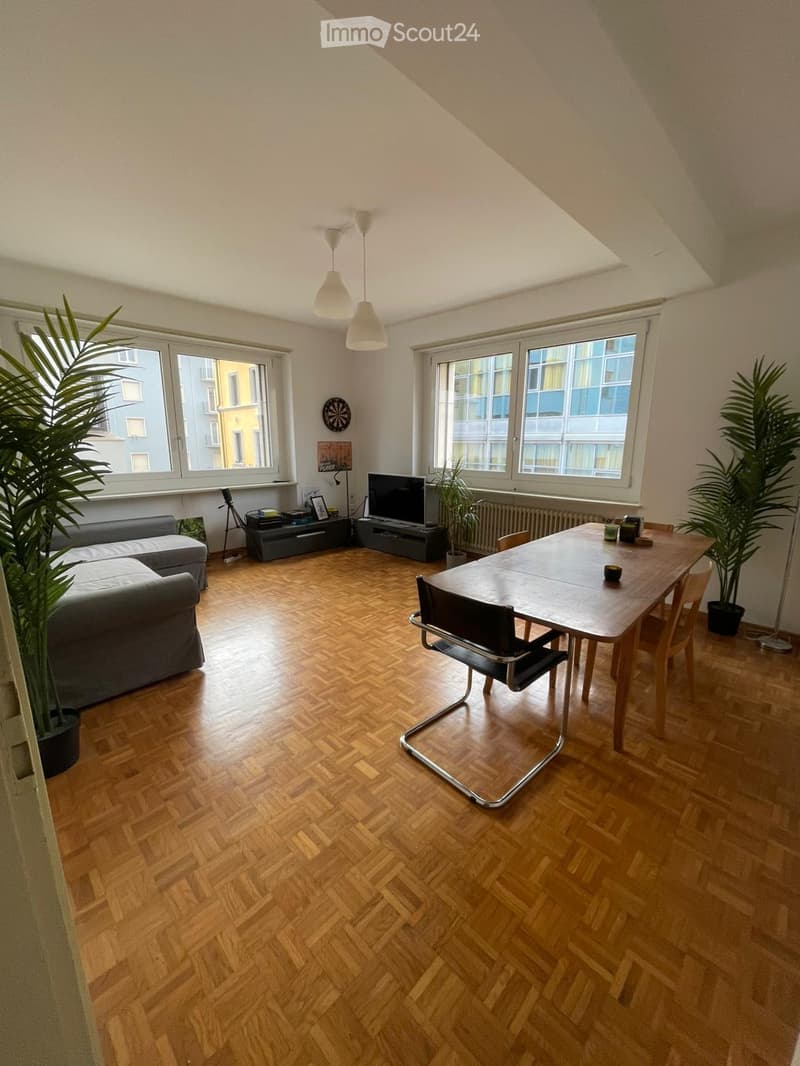 Bright living and dining area with wooden herringbone flooring, large windows, a grey sectional sofa, a wooden dining table with chairs, a television, pendant lights, and plants.