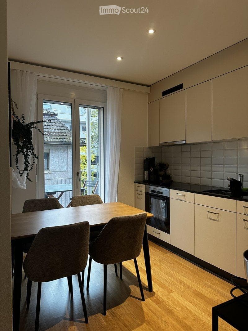 Modern kitchen with light-colored cabinets, a wooden dining table with four chairs, and a coffee maker on the counter. The room features large windows with white curtains and a view of a balcony area.