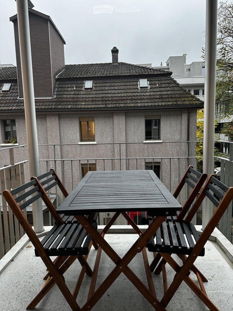 Balcony with a small wooden table and two folding chairs, overlooking a residential building with a tiled roof and multiple windows.