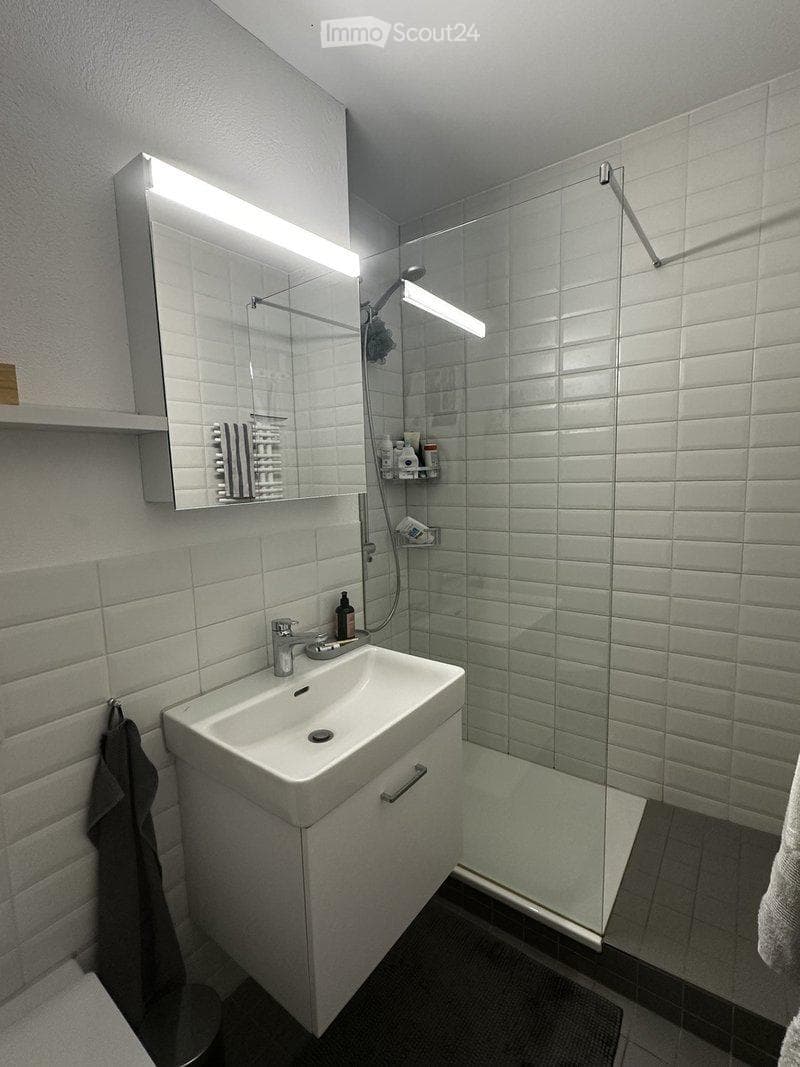 Modern bathroom with white tiled walls, a sink with a mirror, a glass-enclosed shower, and a towel hanging on the wall.