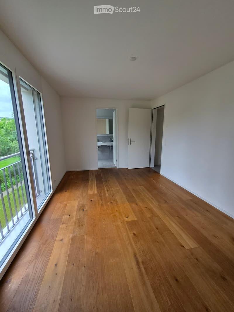 Empty room with wooden flooring, white walls, large window with a view of greenery, and an open doorway leading to a bathroom.