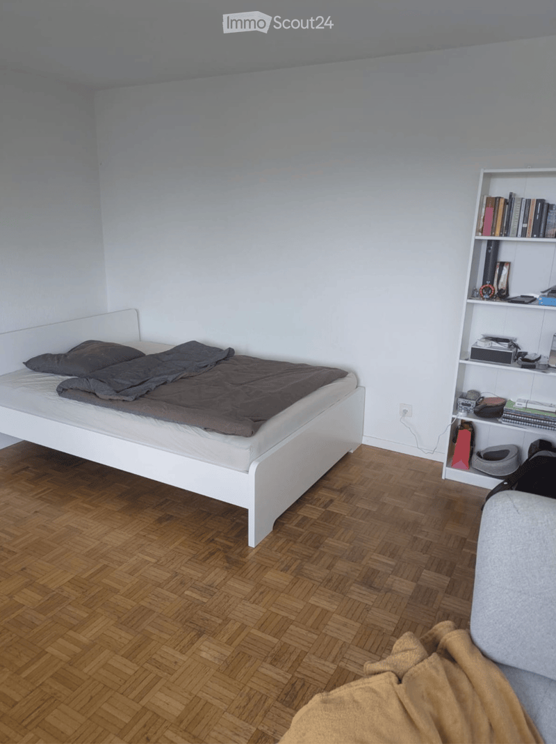 A small bedroom with a white bed, a wooden floor, and a shelf with books and a cat bowl.