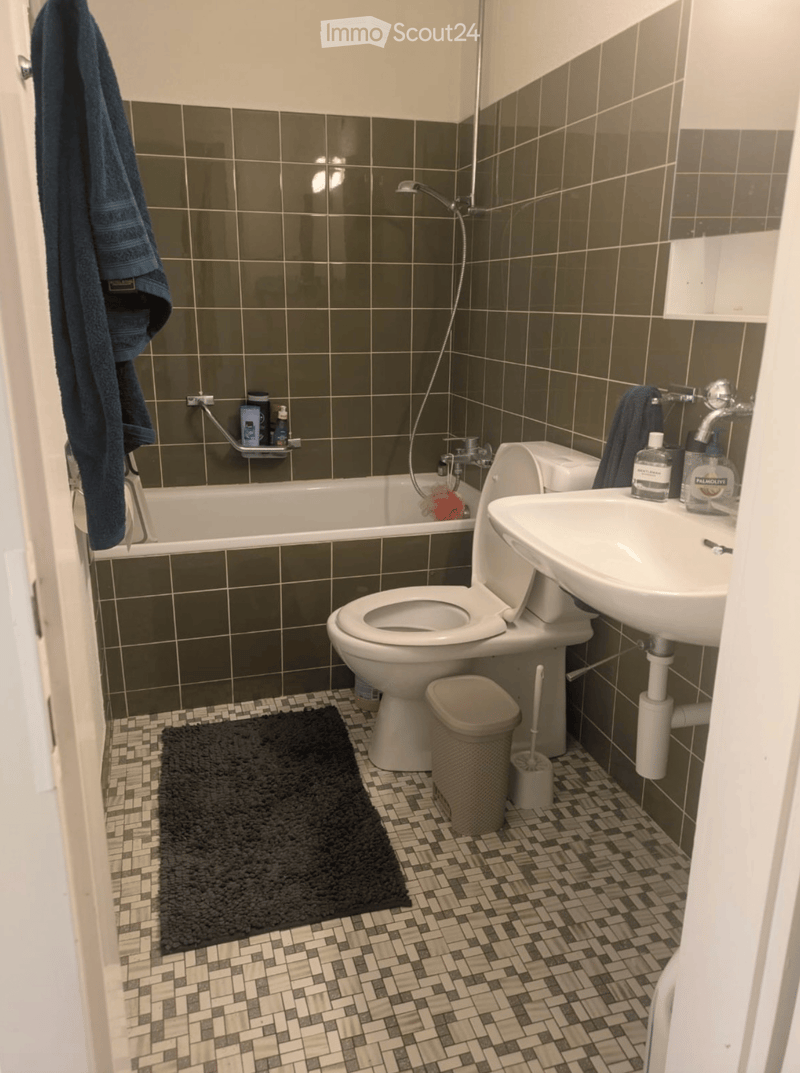 Bathroom with dark green tiled walls, white fixtures, and a black bath mat on the floor.