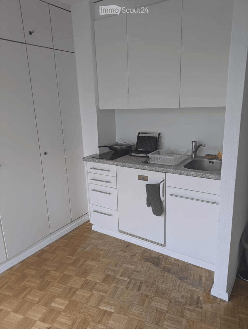Modern kitchen with white cabinets, wooden herringbone flooring, and stainless steel appliances including a sink, oven, and pan on the counter.