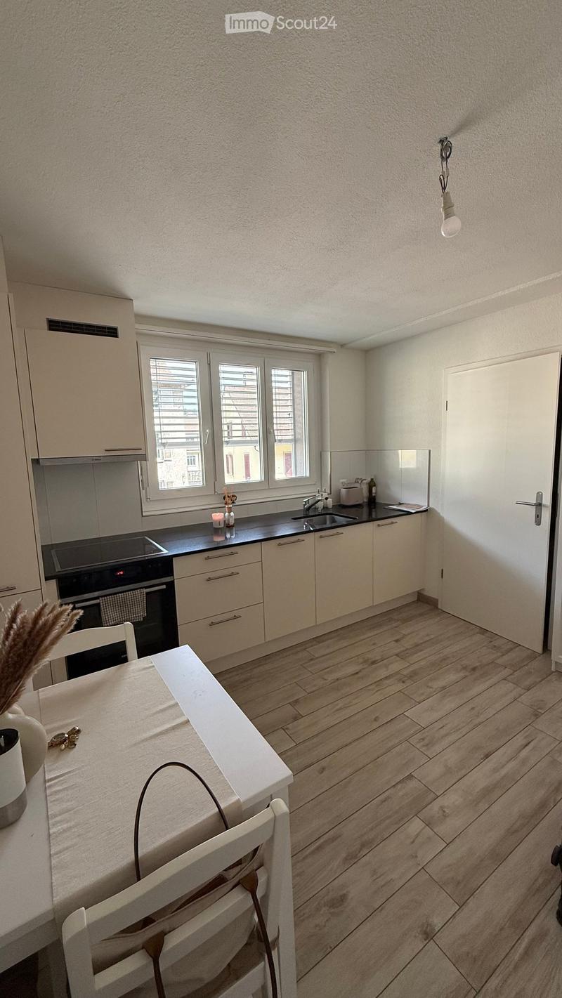 Modern kitchen with light-colored cabinets, a black countertop, and a white dining table with chairs. The floor is wooden, and there is a window with blinds.