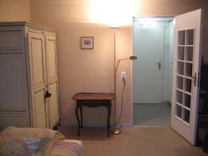 Bedroom with a bed, small wooden table, wardrobe, floor lamp, and a picture frame on the wall. An open door leads to another room.