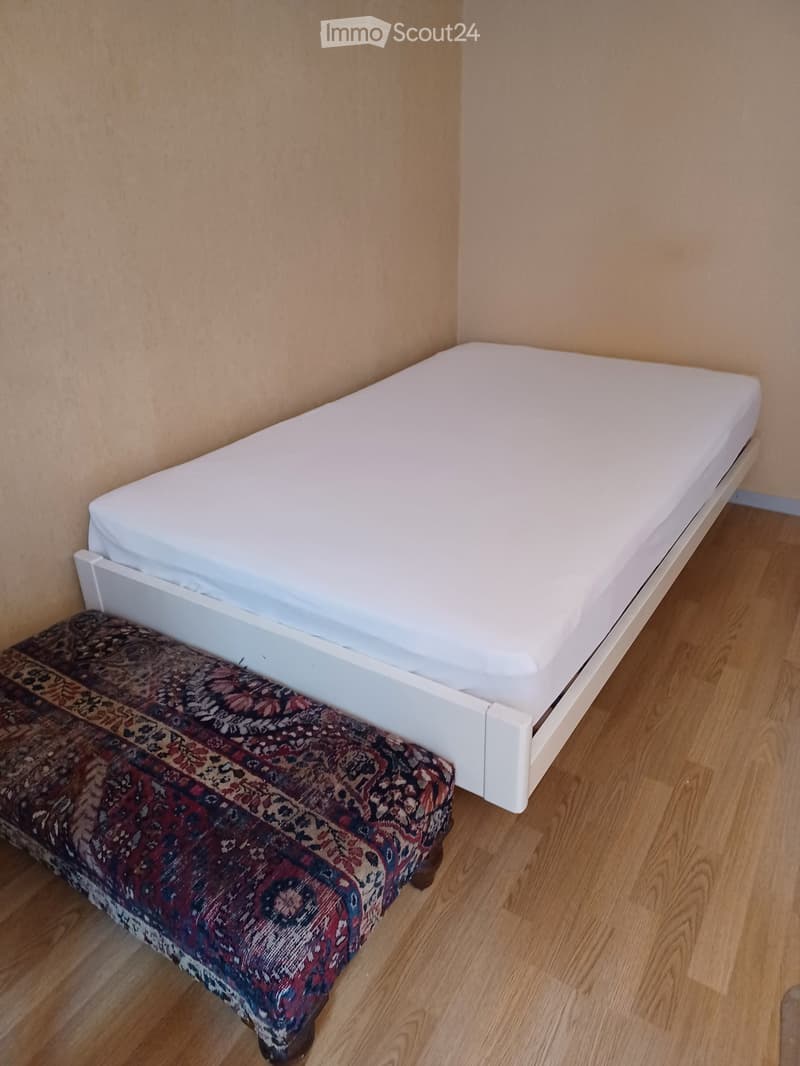 A small, furnished room with a single bed and a patterned bench at the foot of the bed. The room has light wooden flooring and beige walls.