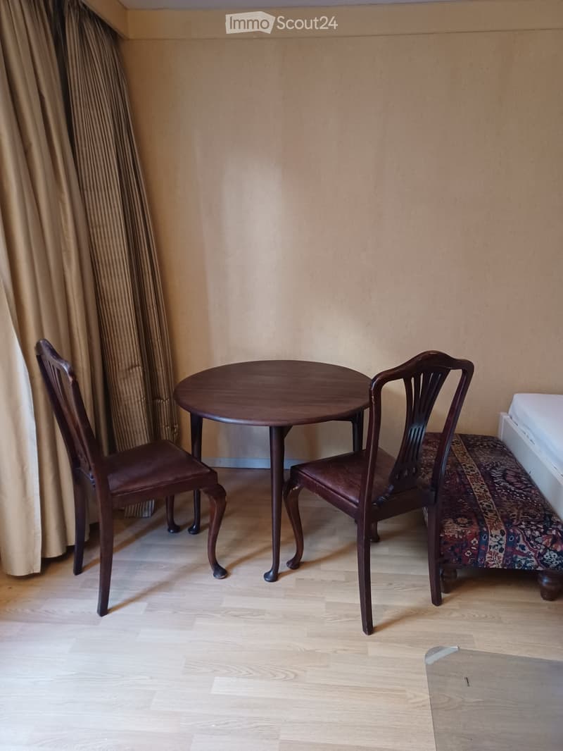 A small, furnished room with a round wooden table and two matching chairs, light wooden flooring, and beige walls.