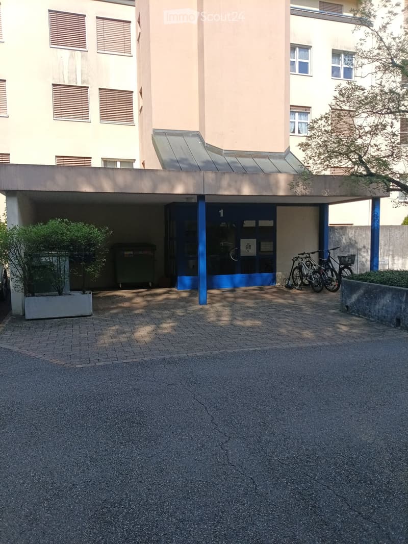 Multi-story building with a covered entrance area, bicycles parked nearby, and a small landscaped area with a tree and planter.