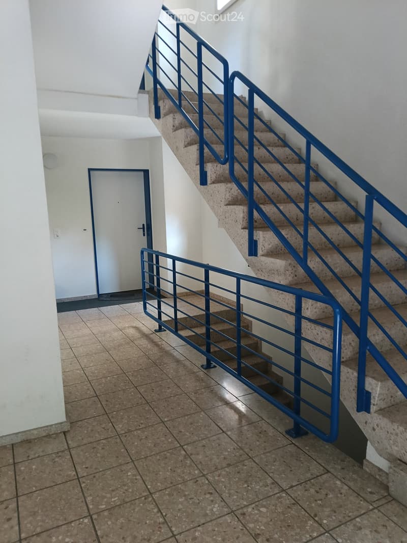 Staircase with blue metal railings and beige tiled steps, leading to an upper level. The hallway has tiled flooring and white walls, with a closed door visible on the left side.