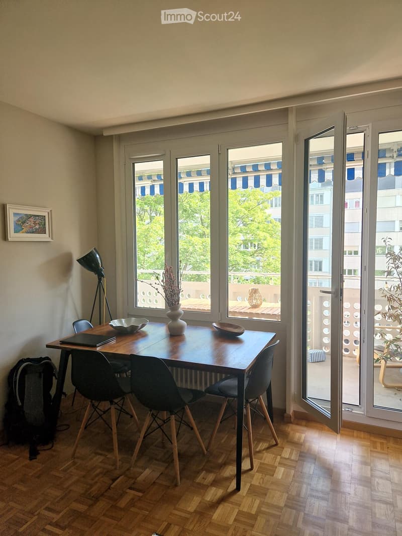 Dining area with a wooden table, four chairs, a vase with dried flowers, a lamp, and a backpack on the floor. Large windows open to a balcony with outdoor furniture.
