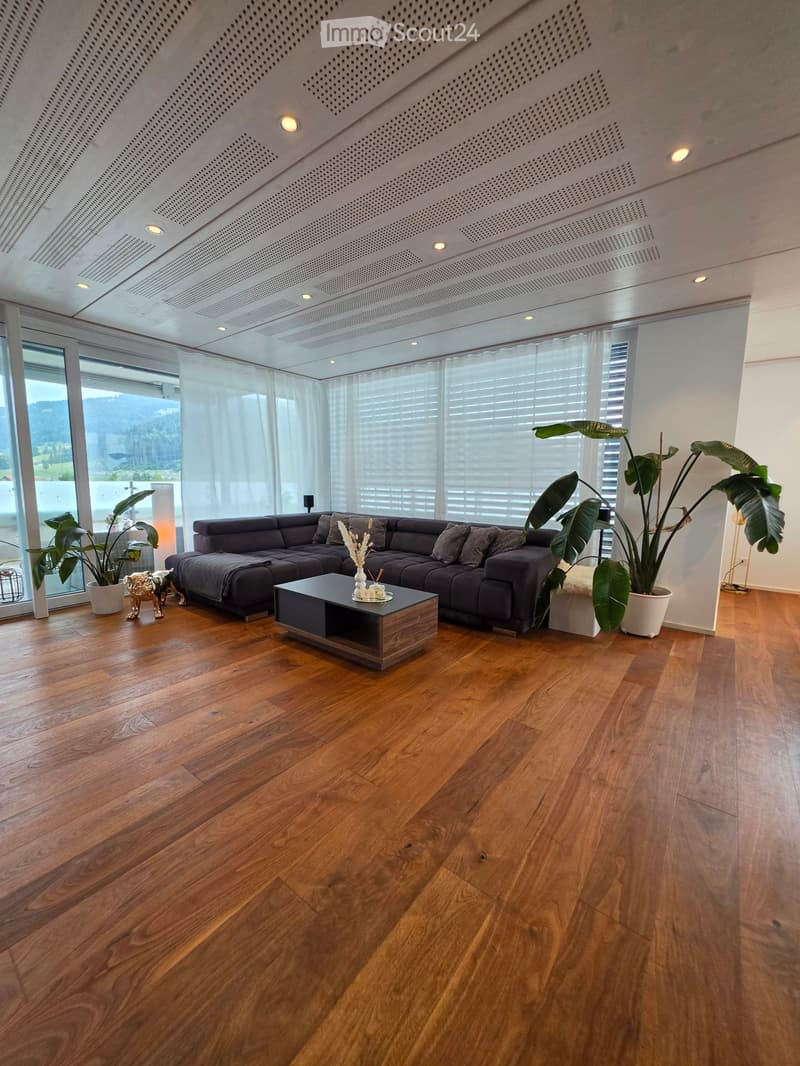 Modern living room with wooden flooring, large windows, grey sectional sofa, coffee table, and potted plants.