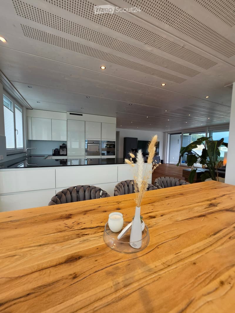 Modern dining area with a wooden dining table, grey upholstered chairs, a vase with dried flowers, and a candle on the table. The background shows a sleek kitchen with white cabinets, stainless steel appliances, and a large window.