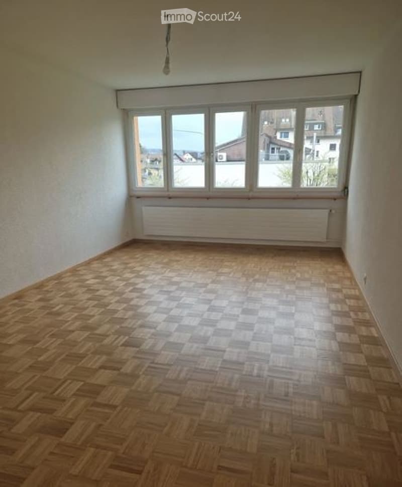 Empty room with wooden parquet flooring, white walls, large windows, and a radiator below the windows.