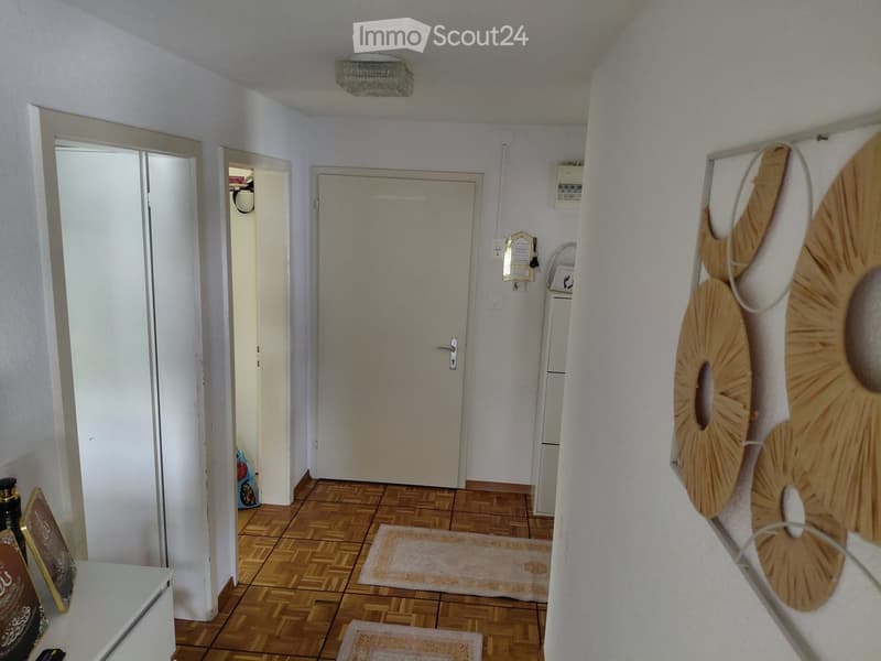 A narrow hallway with wooden flooring, white walls, and several doors leading to different rooms. A large decorative wall piece is visible on the right side.