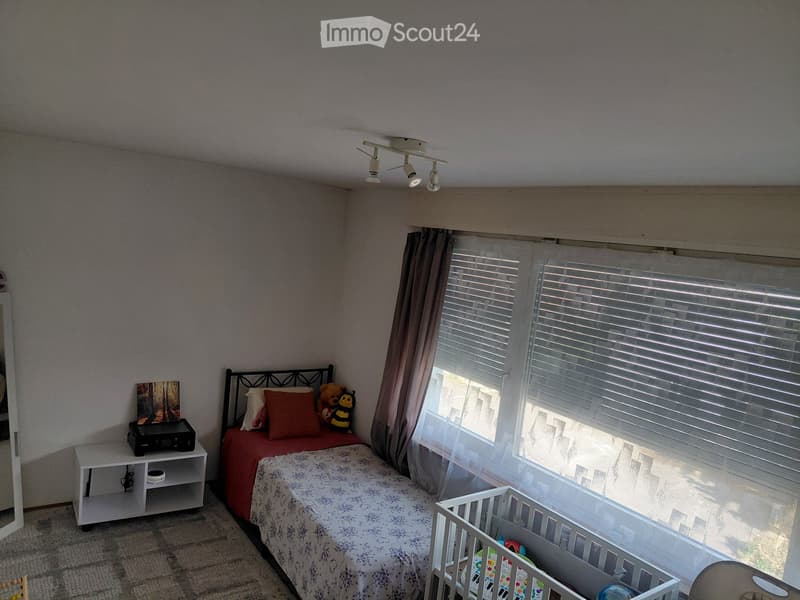 A cozy bedroom with a bed, nightstand, mirror, and a small cradle. The room features large windows with blinds and curtains, and a picture frame on the nightstand.