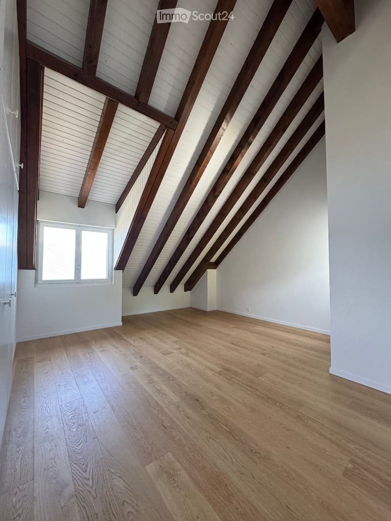 Empty attic space with exposed wooden beams, light wooden flooring, and a small window letting in natural light.