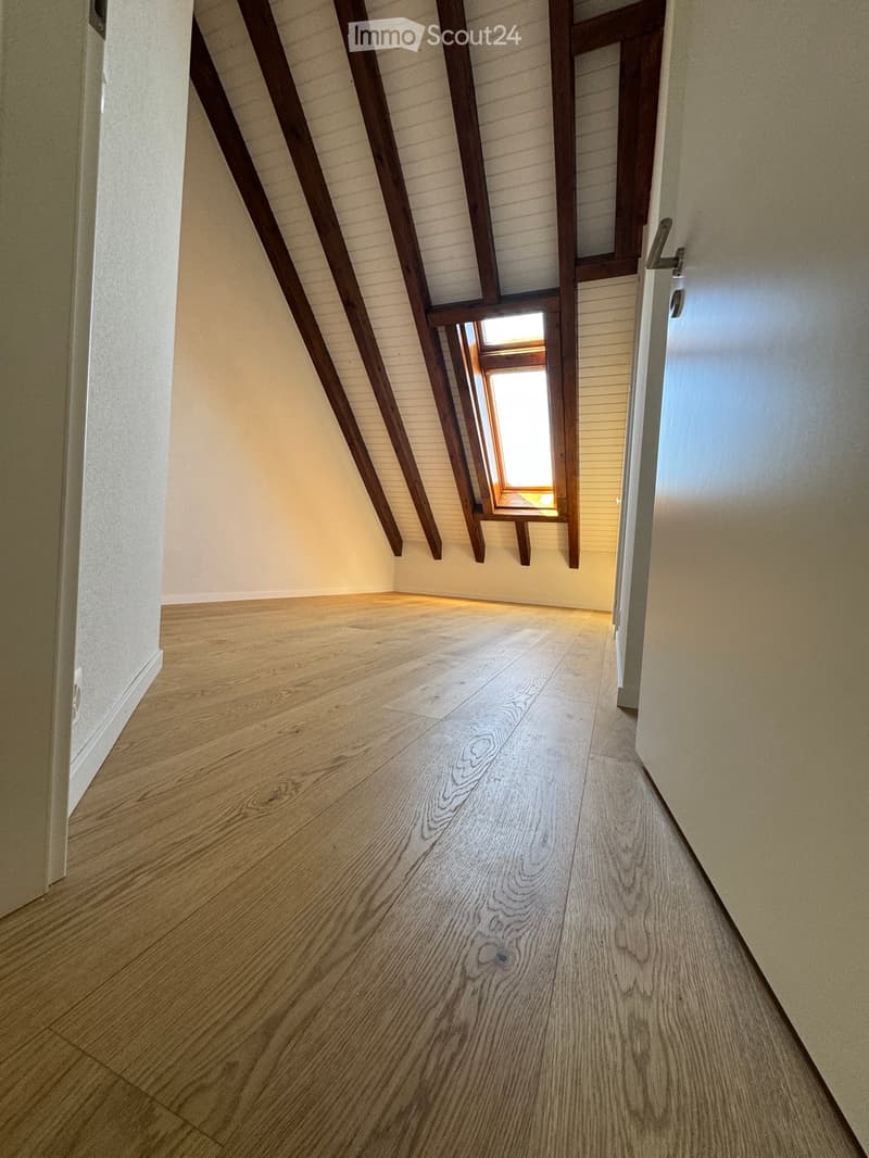 Empty hallway with wooden flooring, white walls, and exposed wooden beams on the ceiling. A small window allows natural light to enter.