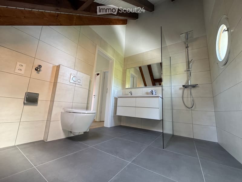 Modern bathroom with a toilet, sink, mirror, and glass-enclosed shower. Light-colored tiles and a dark grey floor.