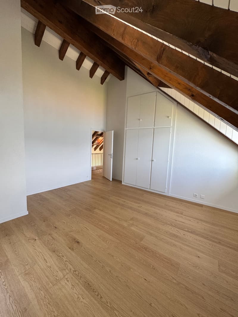 Empty room with light wooden flooring, white walls, exposed wooden beams on the ceiling, and built-in white cabinets.